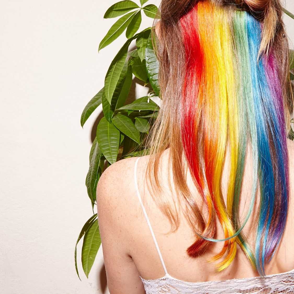 Person with rainbow-colored hair standing against a wall with foliage.