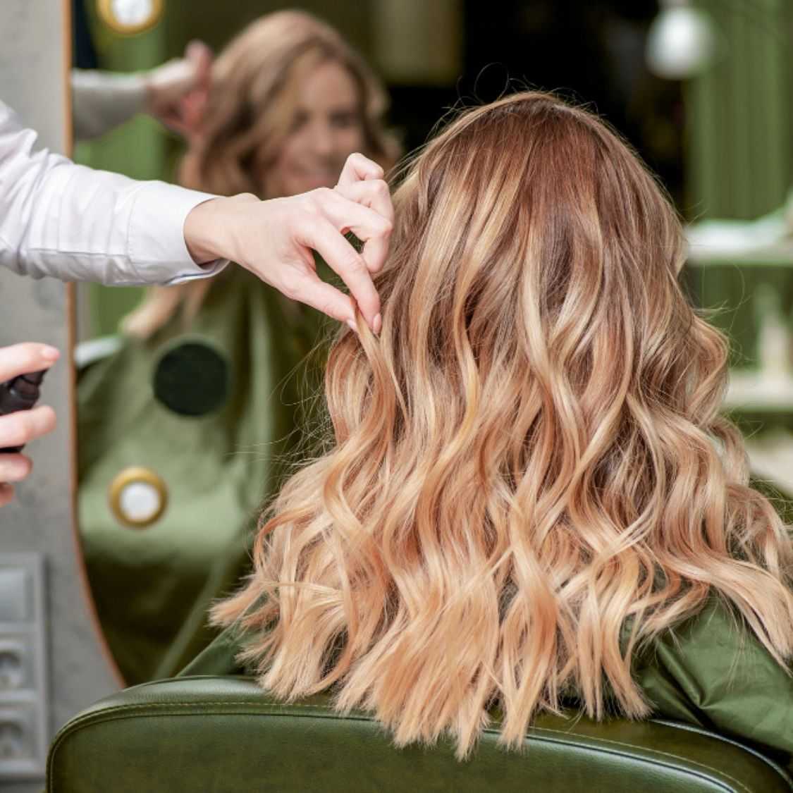 Stylist snipping woman's wavy hair in salon, reflection in mirror.