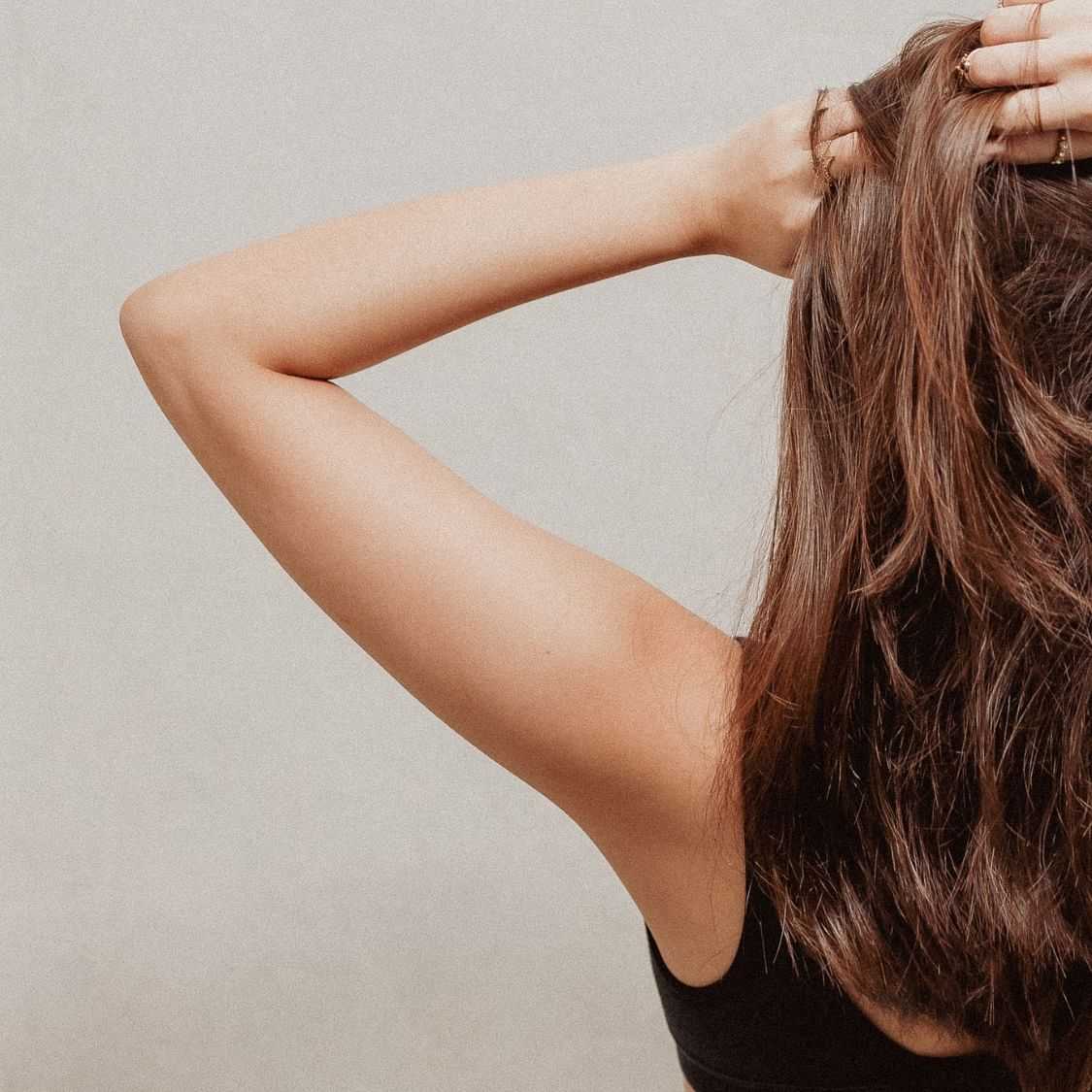 Woman running fingers through her hair, back turned, neutral backdrop.